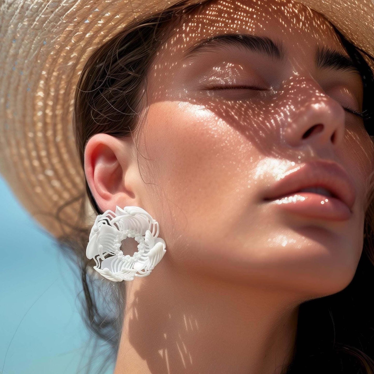 A woman in a straw hat on the beach, demonstrating a large white 3D printed stud earring with a layered floral-geometric patt