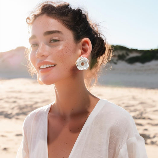 A smiling woman on a beach, wearing a white top, demonstrating a large white 3D printed stud earring with an intricate geometric pattern.