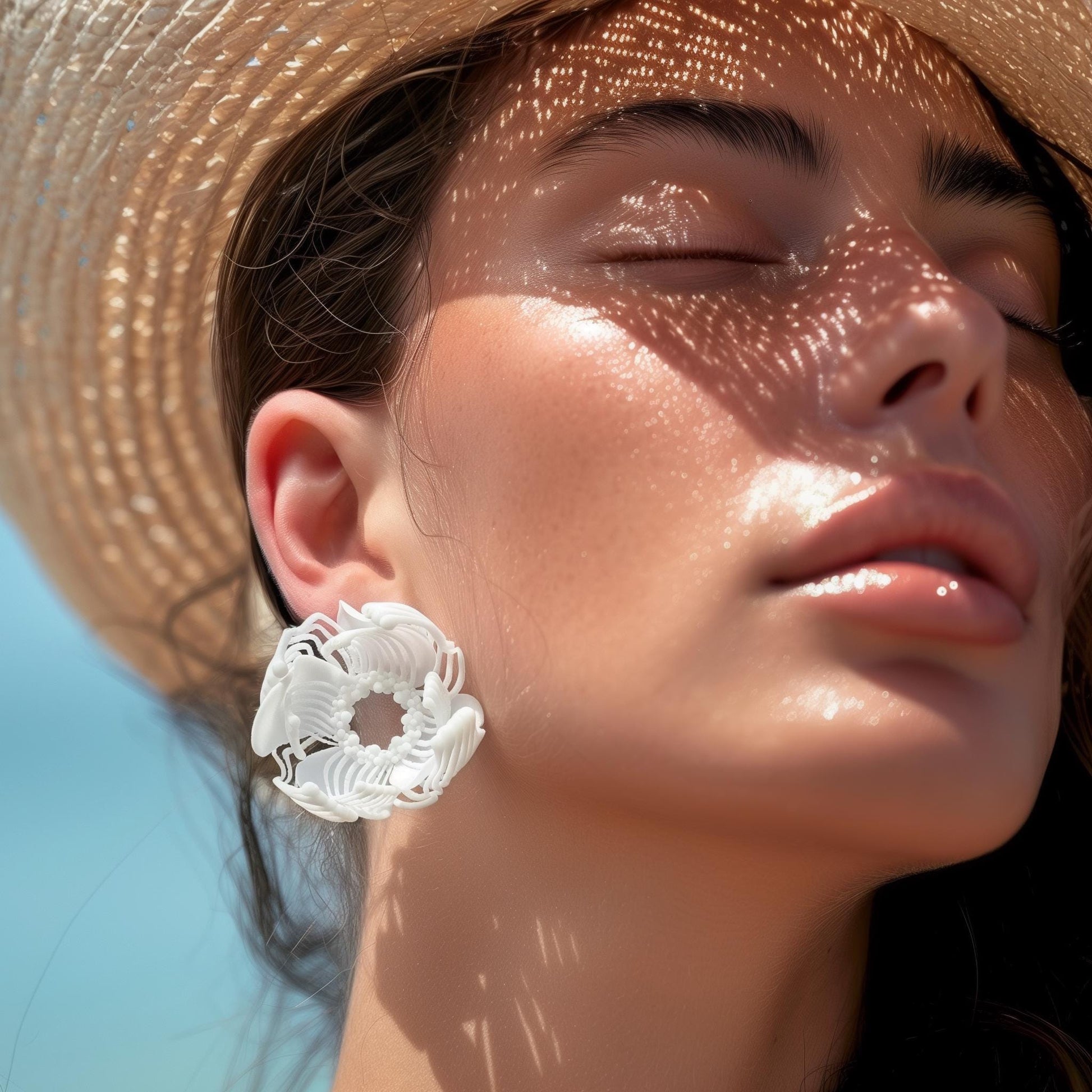 A woman in a straw hat on the beach, demonstrating a large white 3D printed stud earring with a layered floral-geometric pattern.