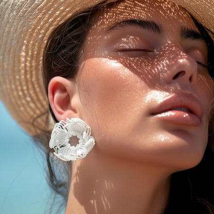 A woman in a straw hat on the beach, demonstrating a large white 3D printed stud earring with a layered floral-geometric pattern.