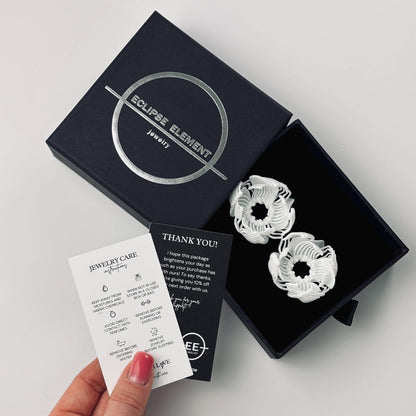 A hand holding brand cards and a booklet, next to black and white 3D printed stud earrings with a complex floral-geometric design.