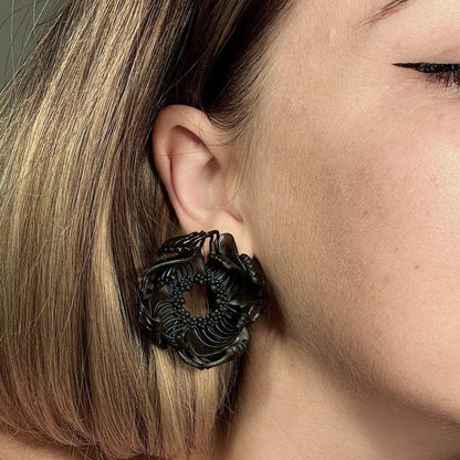 A close-up of a woman's face, demonstrating a pair of black 3D printed stud earrings with a detailed, interlocking geometric pattern.