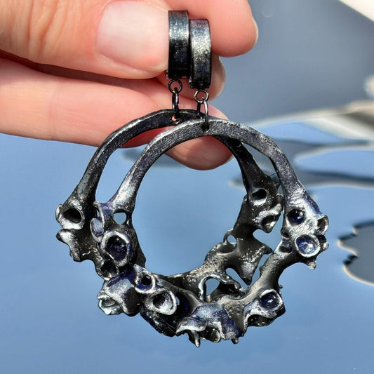 Black statement earring with a geometric hoop design and what appears to be custom metallic accents, held in a hand with a reflective background.