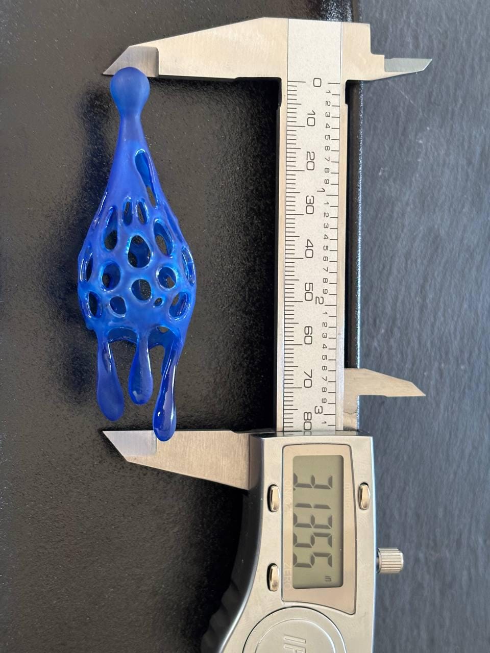 Blue 3D-printed earrings being measured with a digital caliper on a dark background.