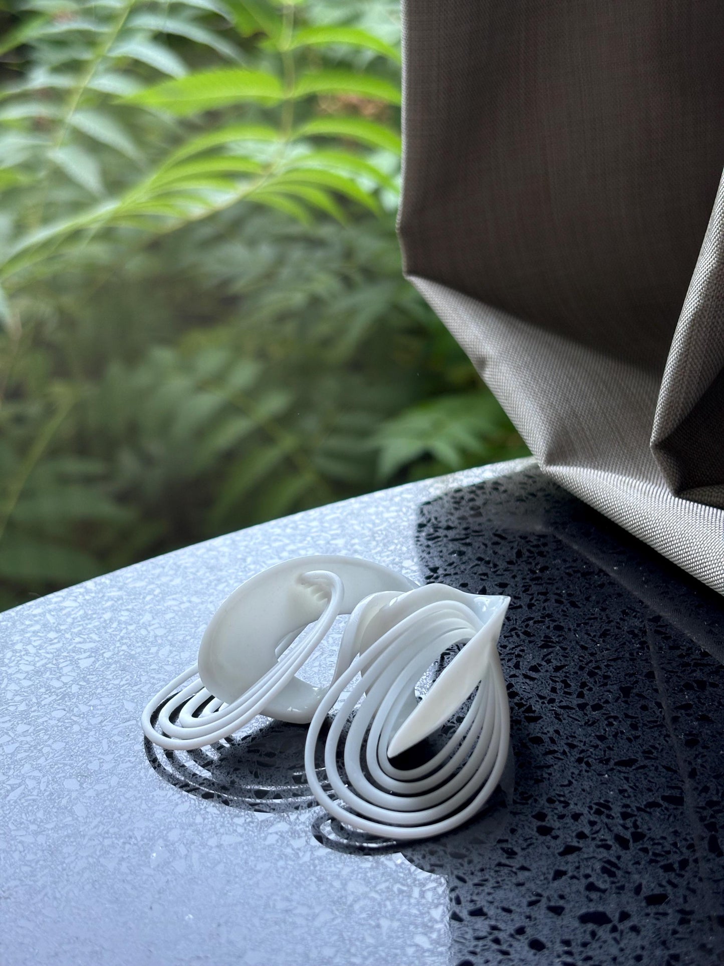 White Infinity Loops Statement Earrings on a wet surface with blurred green foliage in the background.
