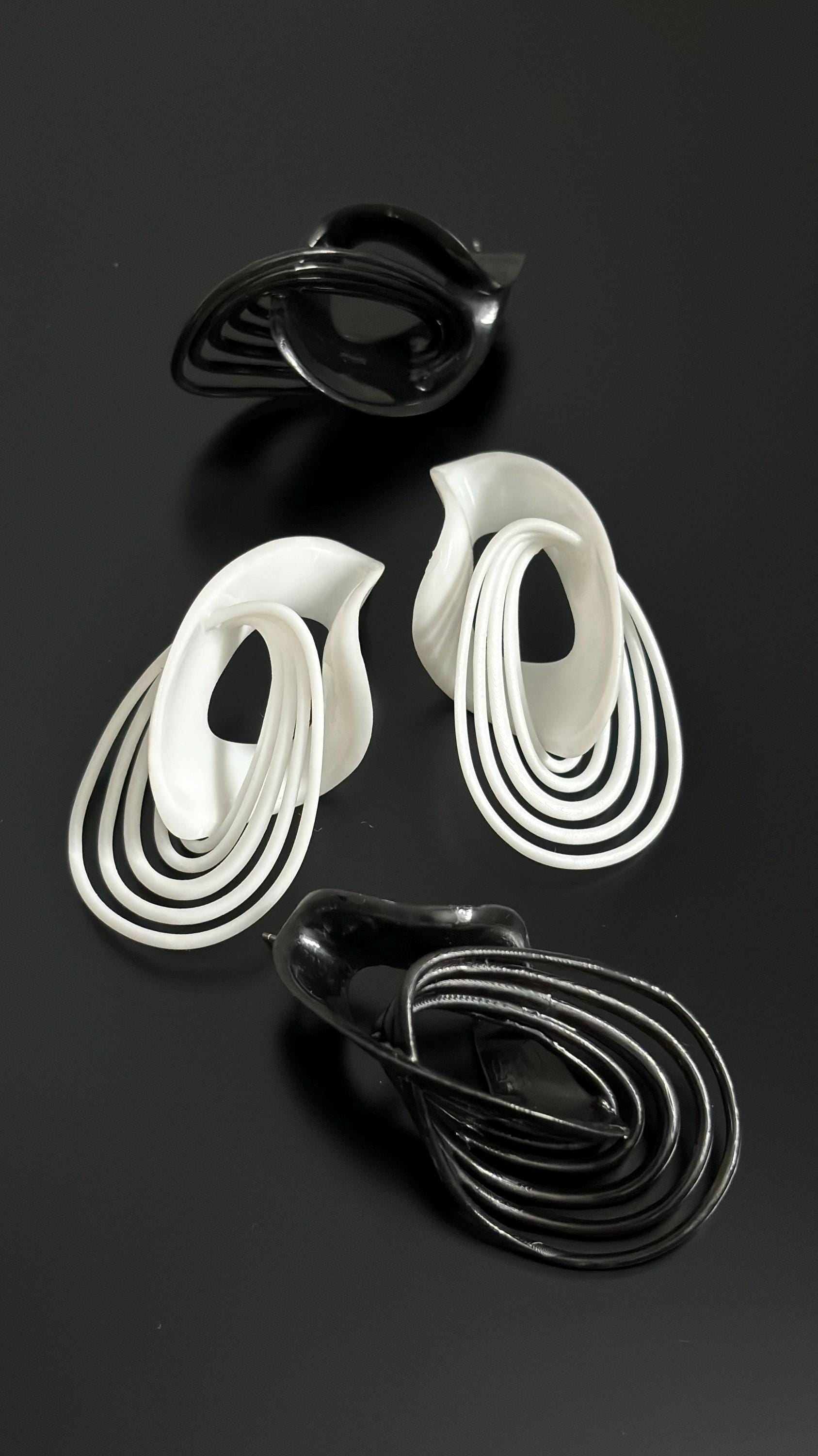 A pair of sculptural drop earrings in white and black, with a fluid, architectural design, displayed against a dark background.