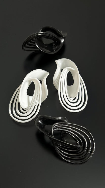 A pair of sculptural drop earrings in white and black, with a fluid, architectural design, displayed against a dark background.