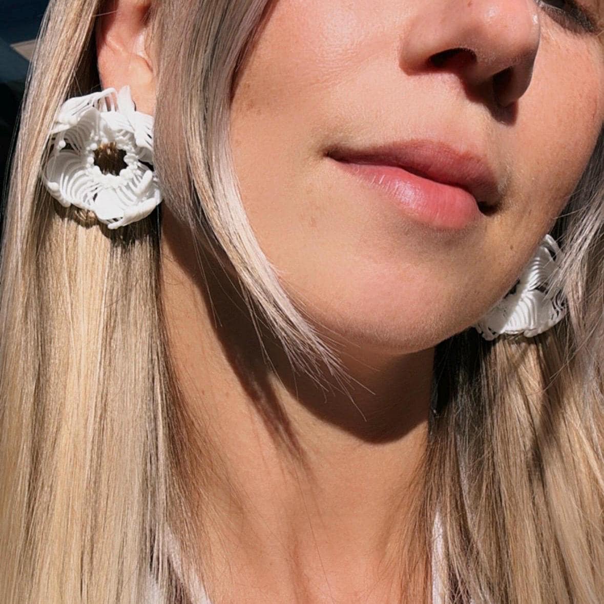 A close-up of a woman's face, demonstrating a pair of white 3D printed stud earrings with an intricate floral-geometric design.