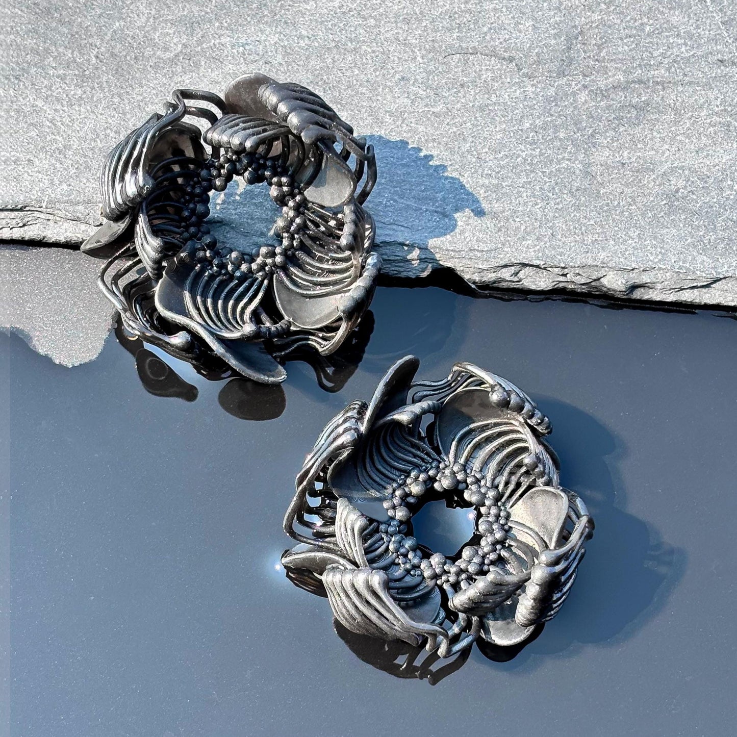 Two black 3D printed stud earrings with an intricate pattern, resting on a grey, rocky surface against a blue background.