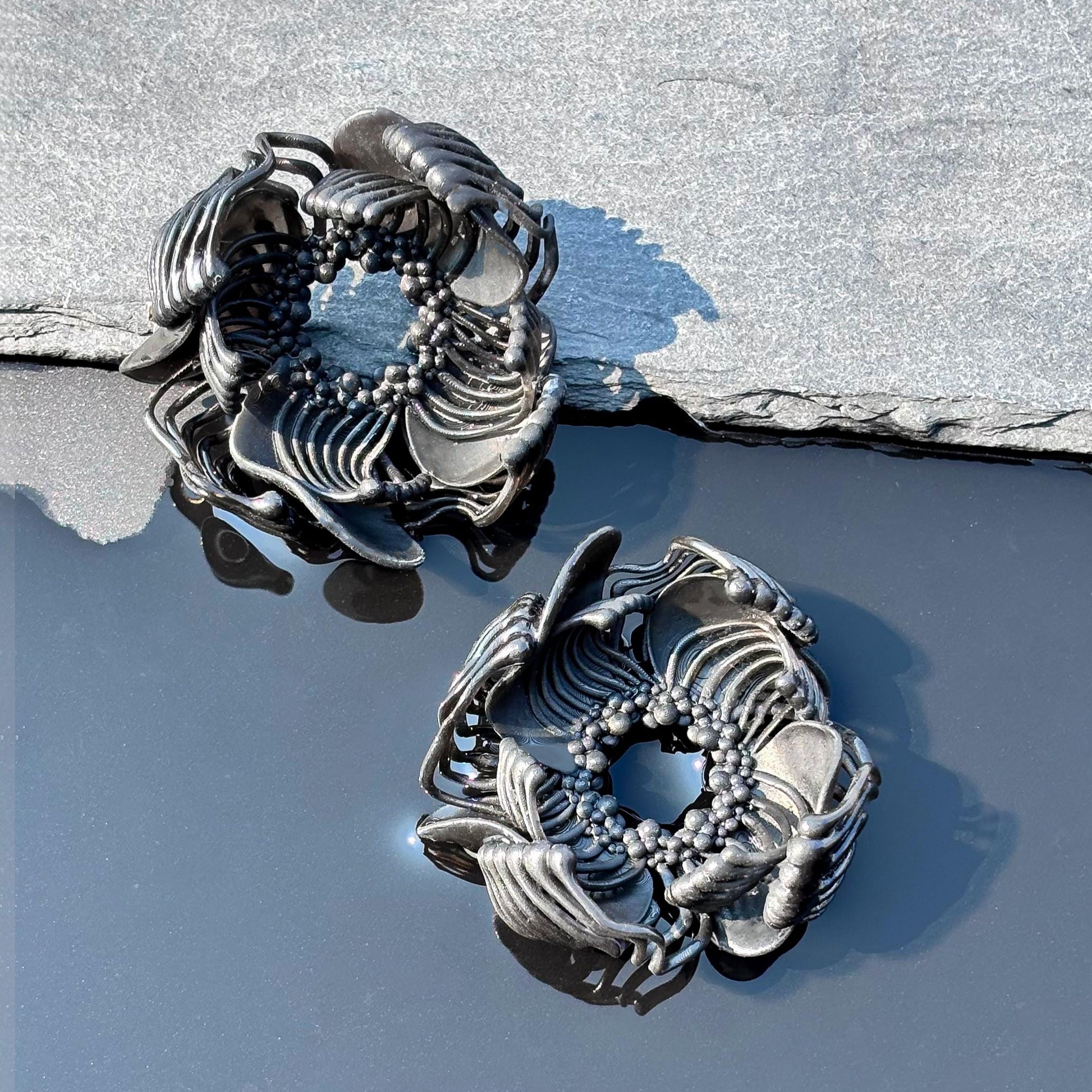 Two black 3D printed stud earrings with an intricate pattern, resting on a grey, rocky surface against a blue background.