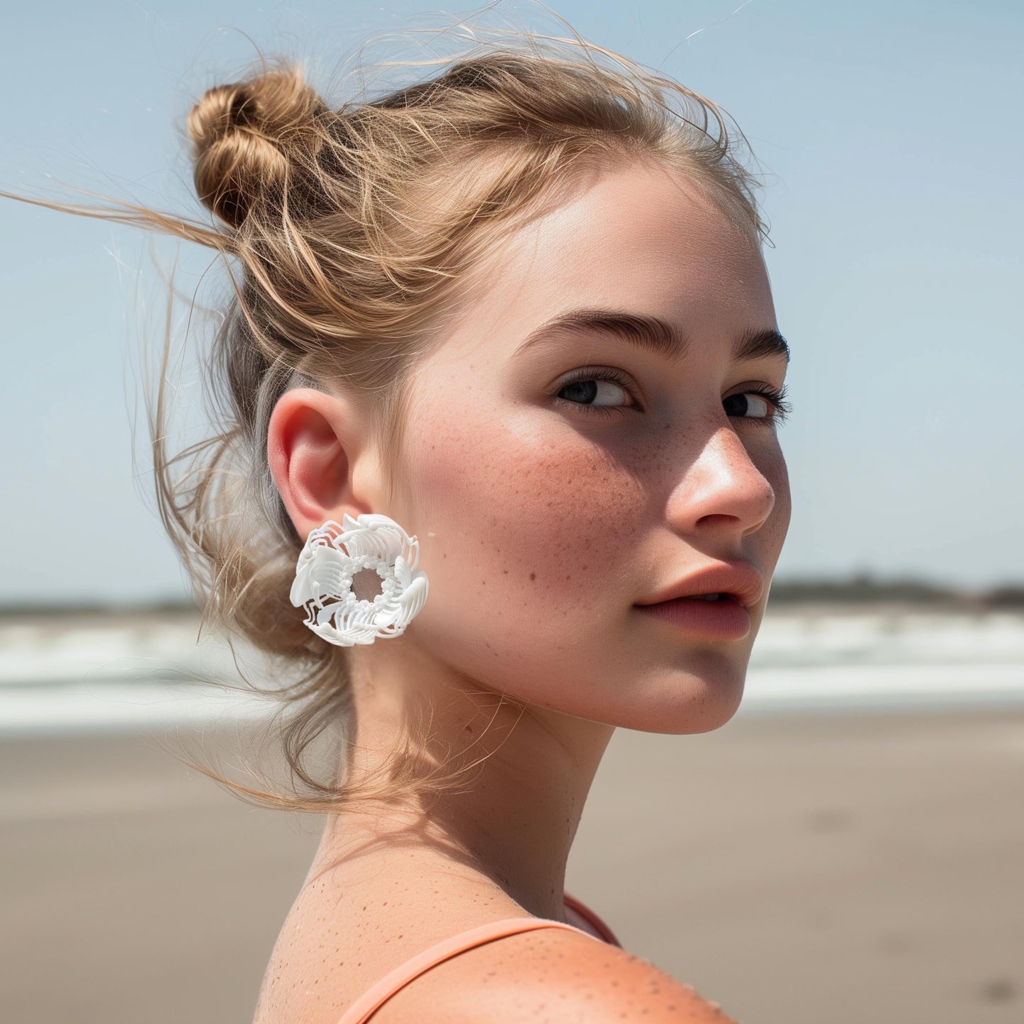 A young woman on a beach, looking away, showcasing a large white 3D printed stud earring with an intricate geometric pattern.