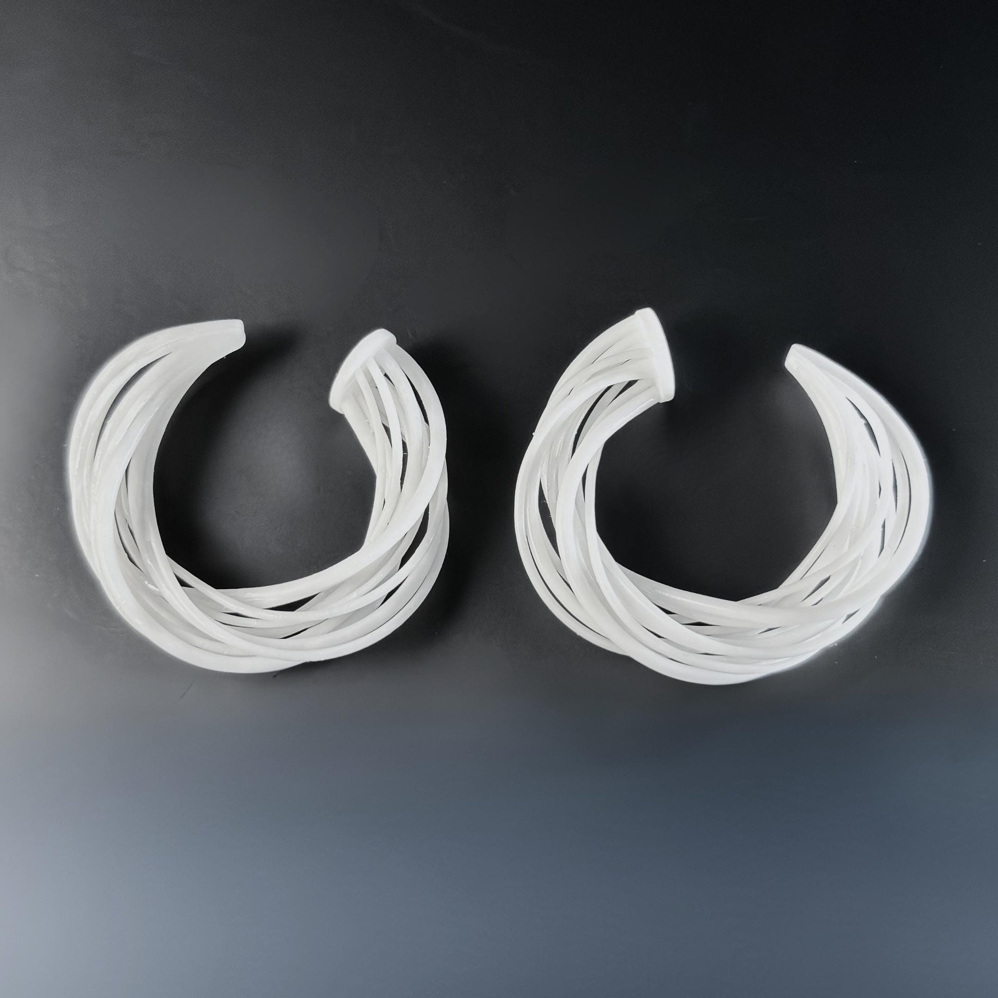 Two large white 3D printed hoop earrings, featuring a complex design of layered, swirling strands, set against a dark, black background.