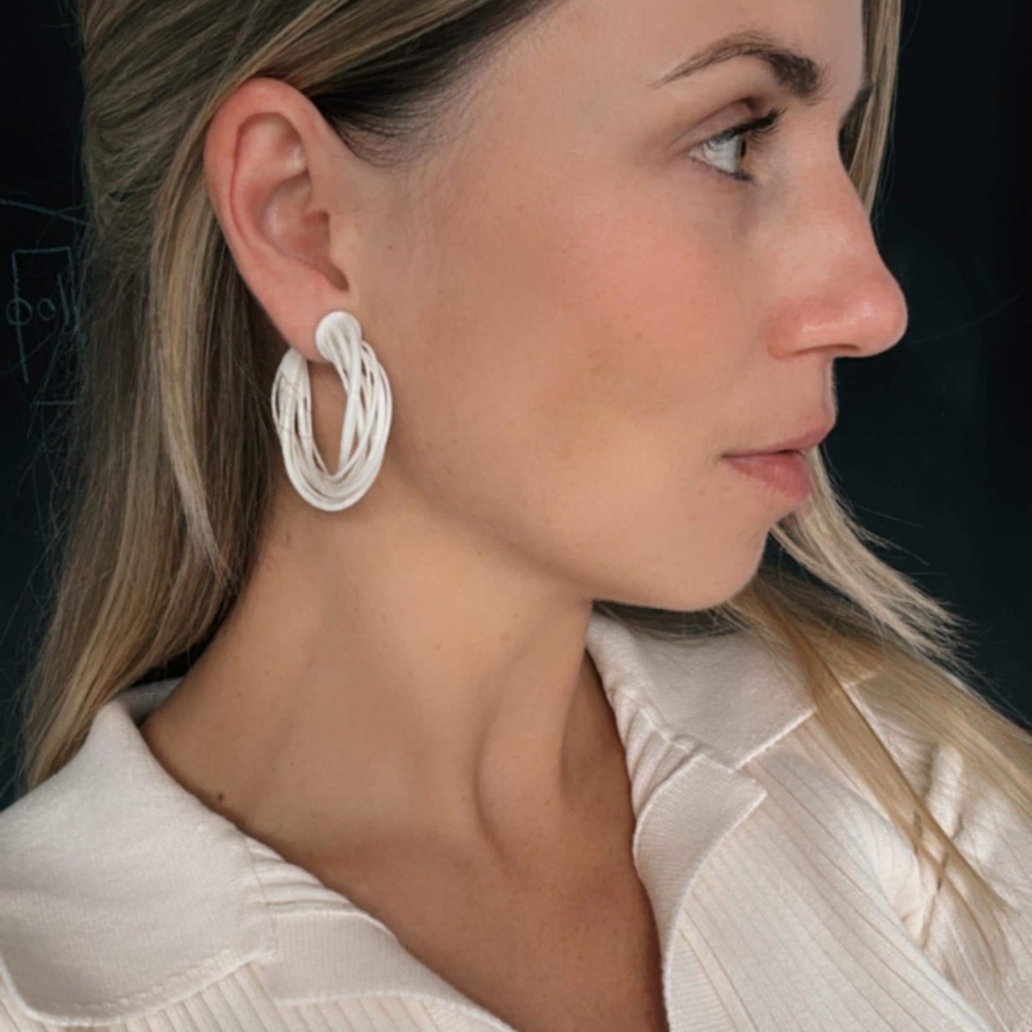 A model wearing a white satin top, viewed in profile, demonstrating a large white 3D printed Swirl Hoop Earring on her ear.