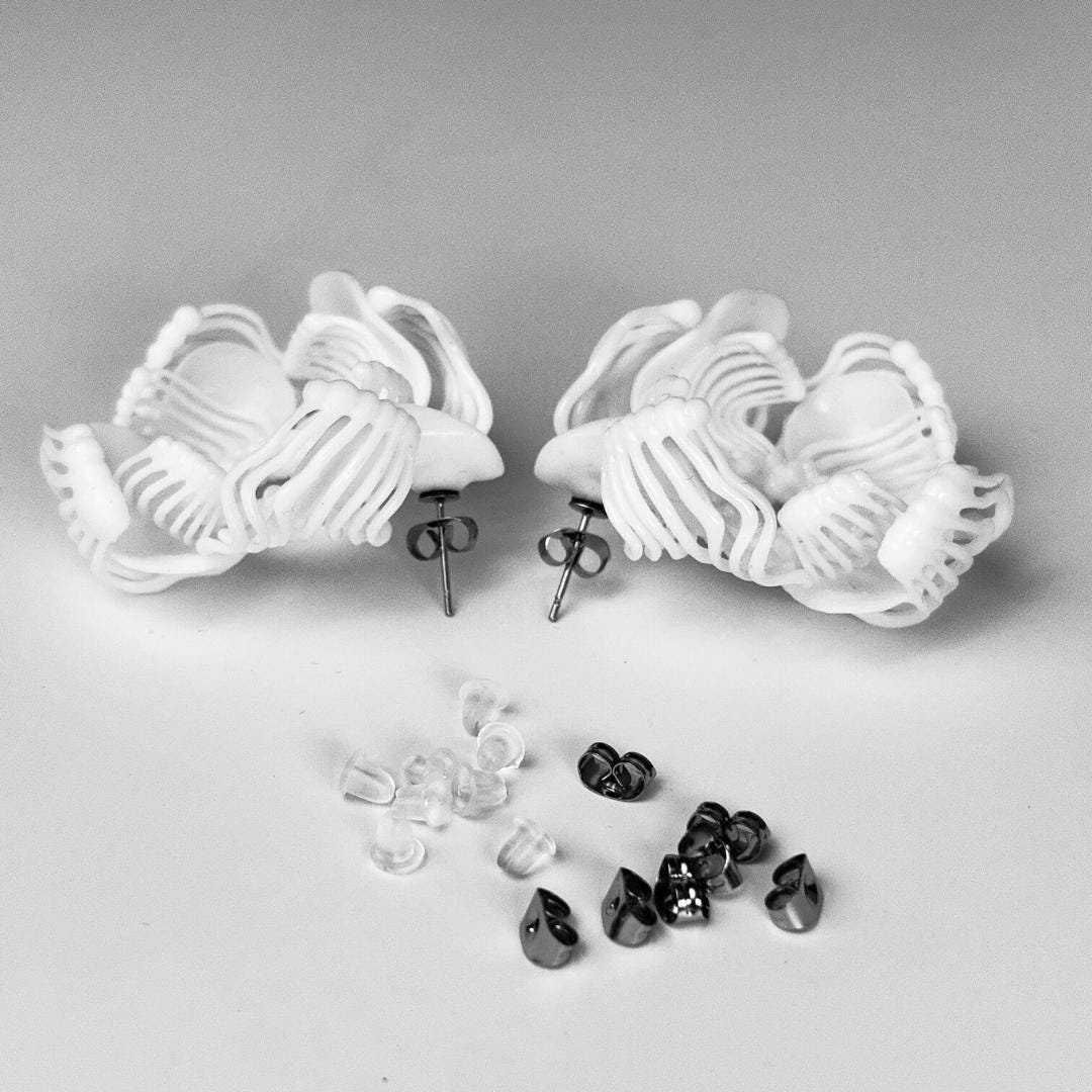 Two white 3D printed stud earrings with a complex design, lying on a white background next to a scattering of black and white beads/pearls.