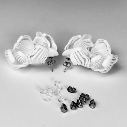 Two white 3D printed stud earrings with a complex design, lying on a white background next to a scattering of black and white beads/pearls.