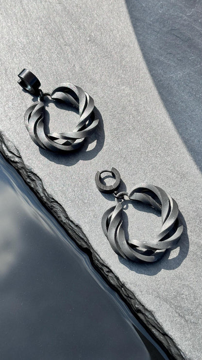 A pair of black, hoop-style statement earrings captured on a reflective surface, likely water, with a bright, sunny background.