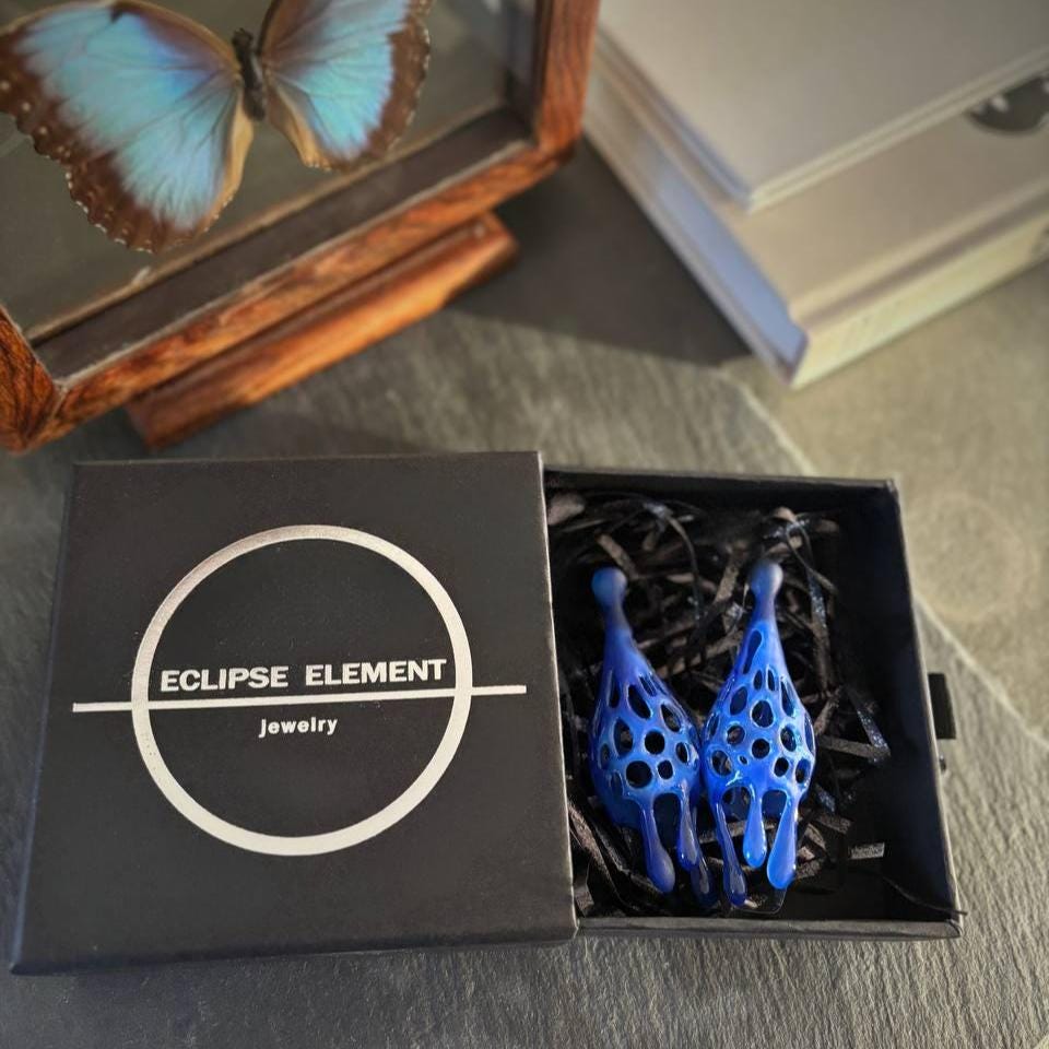 A pair of blue sculptural drop earrings made of 3D printed blue resin, displayed in an open black box with 'ECLIPSE ELEMENT jewelry' printed on the lid.